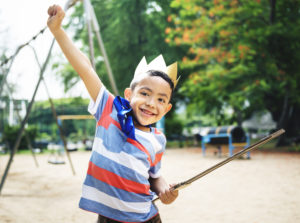 Excited preschool boy on playground wearing crown with cardboard sword